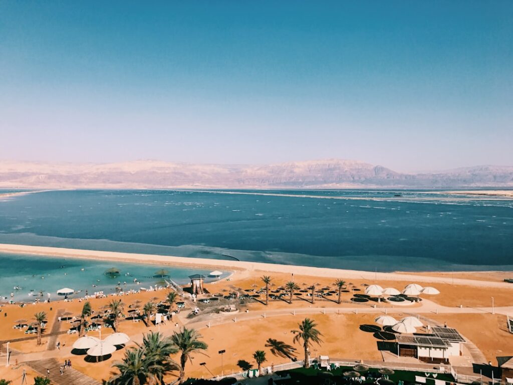 נוף פנורמי של צפון ים המלח עם מים תכולים, הרי מדבר צחיחים ועצי תמר ירוקים באופק