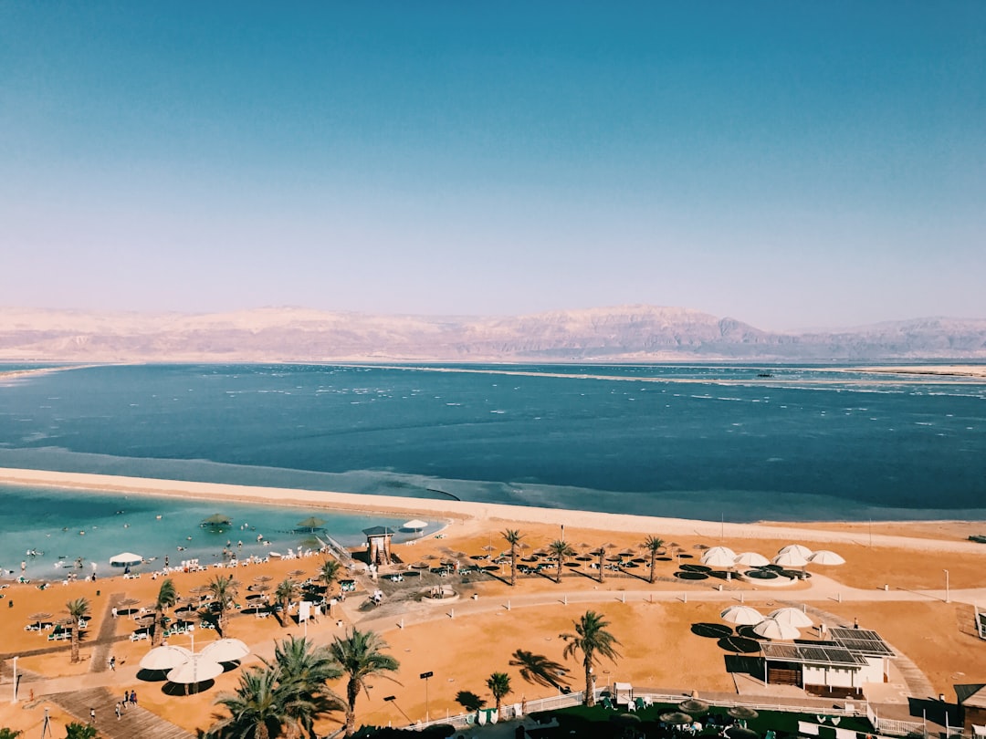 נוף פנורמי של צפון ים המלח עם מים תכולים, הרי מדבר צחיחים ועצי תמר ירוקים באופק