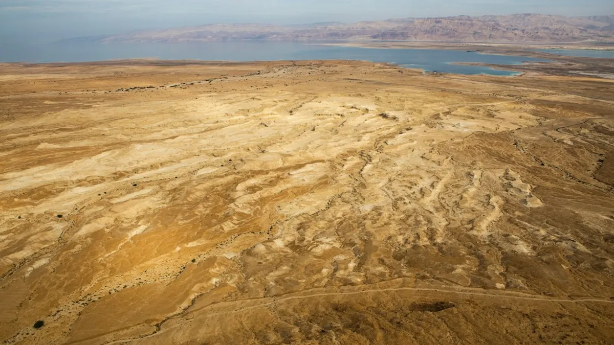 נוף אווירי של ים המלח והמדבר שמסביב, עם גוונים של חום וצהוב באדמה, ומים כחולים-ירוקים של הים ברקע, והרים בצבע חום בהיר באופק