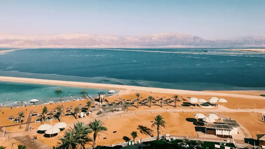 נוף פנורמי של צפון ים המלח עם מים תכולים, הרי מדבר צחיחים ועצי תמר ירוקים באופק